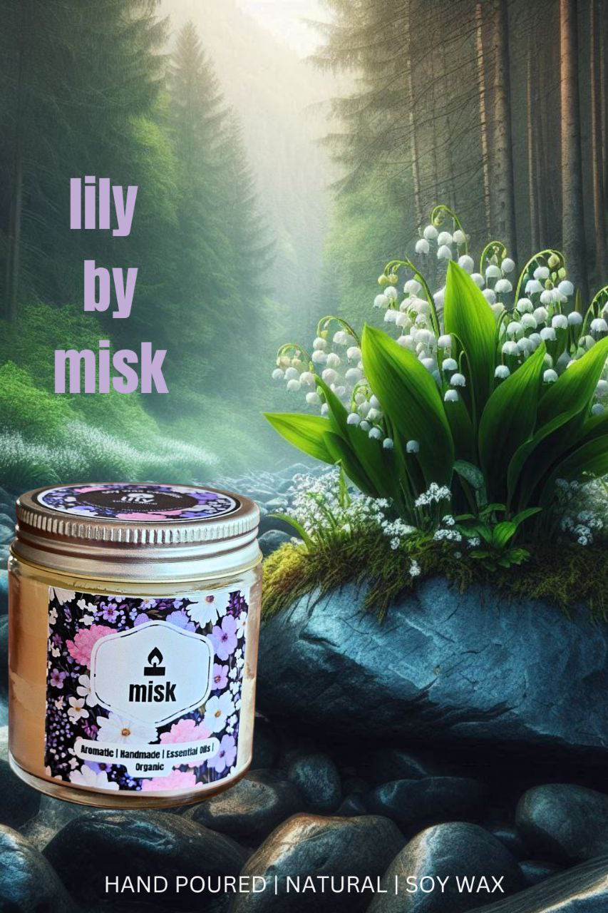 A promotional image for "lily by misk" candles, featuring a jar candle with a floral label and the brand logo "misk." The candle is placed on rocks beside a cluster of white lily-of-the-valley flowers with green leaves. In the background, a misty forest with tall trees is visible. The image also includes text overlays: "lily by misk," "Aromatic | Handmade | Essential oils | Organic," and "HAND POURED | NATURAL | SOY WAX."