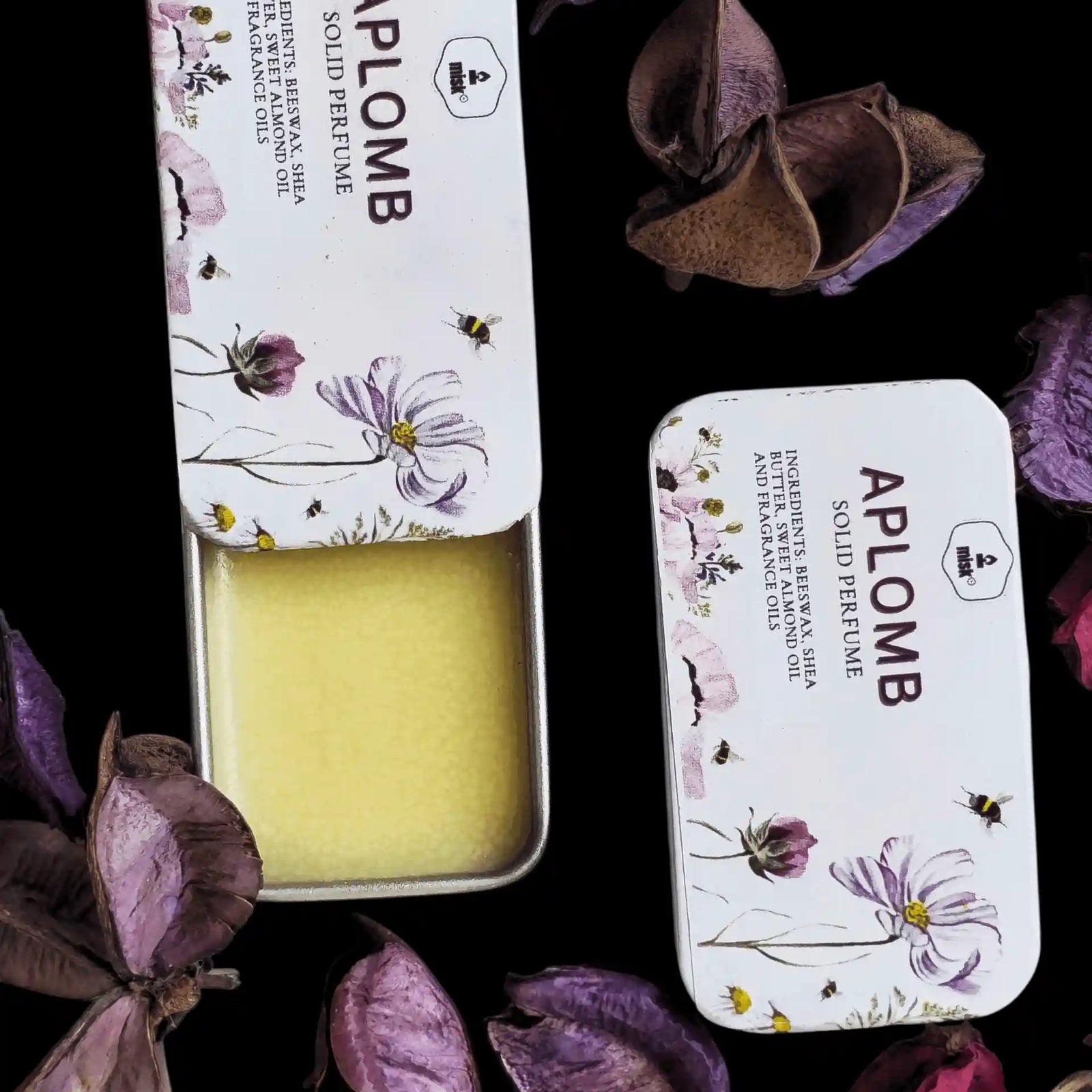 Aplomb solid perfume in a tin surrounded by purple flowers on a dark background