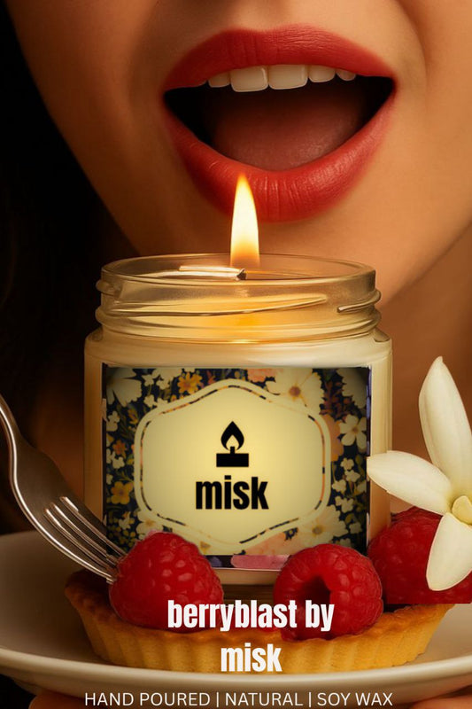 A young woman with red lipstick with mouth open as if to blow out the candle. In the foreground a lit candle in a jar labelled “misk” with a floral pattern and the text “ berryblast by misk” below it. The candle is placed on a small tart with raspberries, and a fork is visible on the left