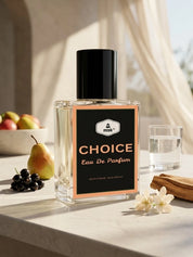 A clear rectangular perfume bottle labeled 'CHOICE Eau De Parfum' with a black cap and black label, styled on a light stone surface with fruit, a glass of water, and flowers in a bright indoor setting.