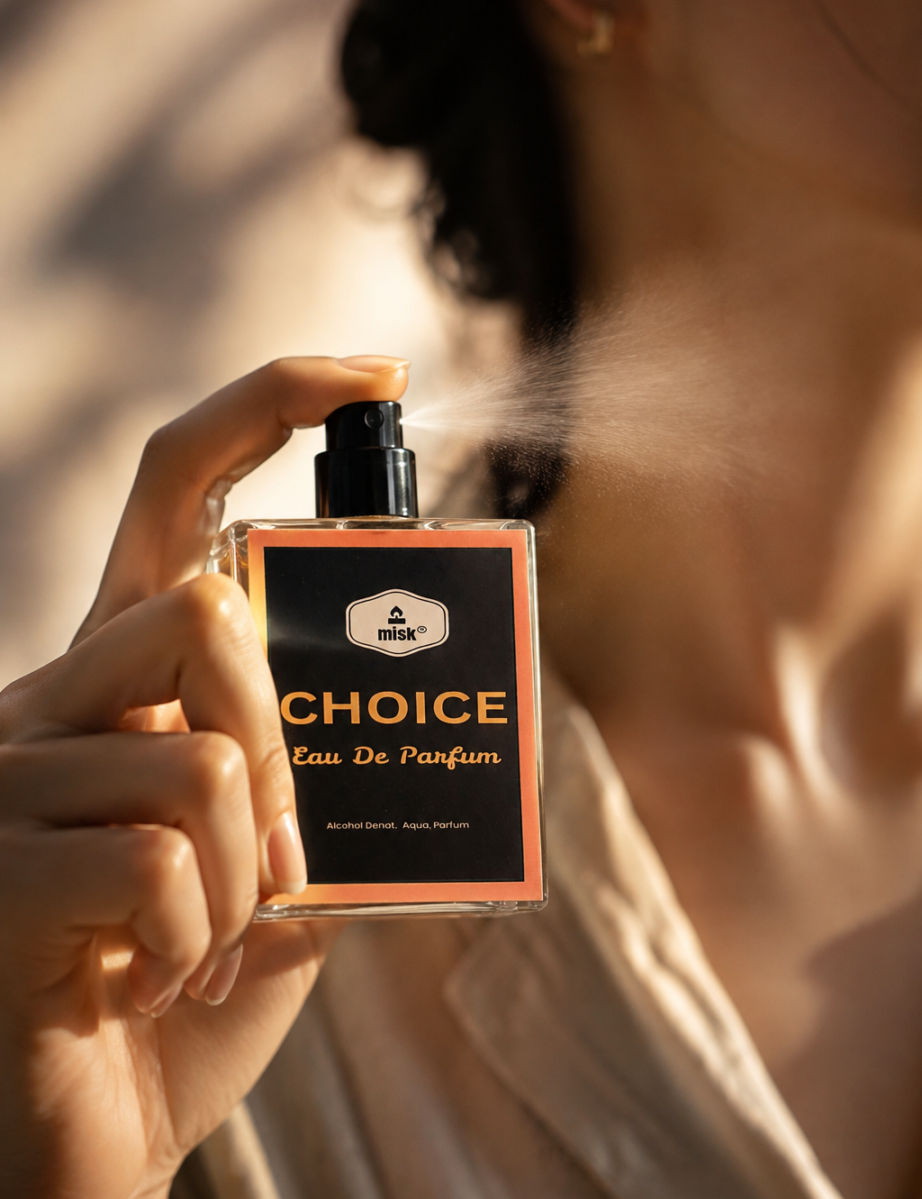 A person holding a bottle of CHOICE Eau De Parfum, spraying a fine mist. The person is wearing a light beige satin shirt, and the background is softly blurred with warm sunlight and shadows.
