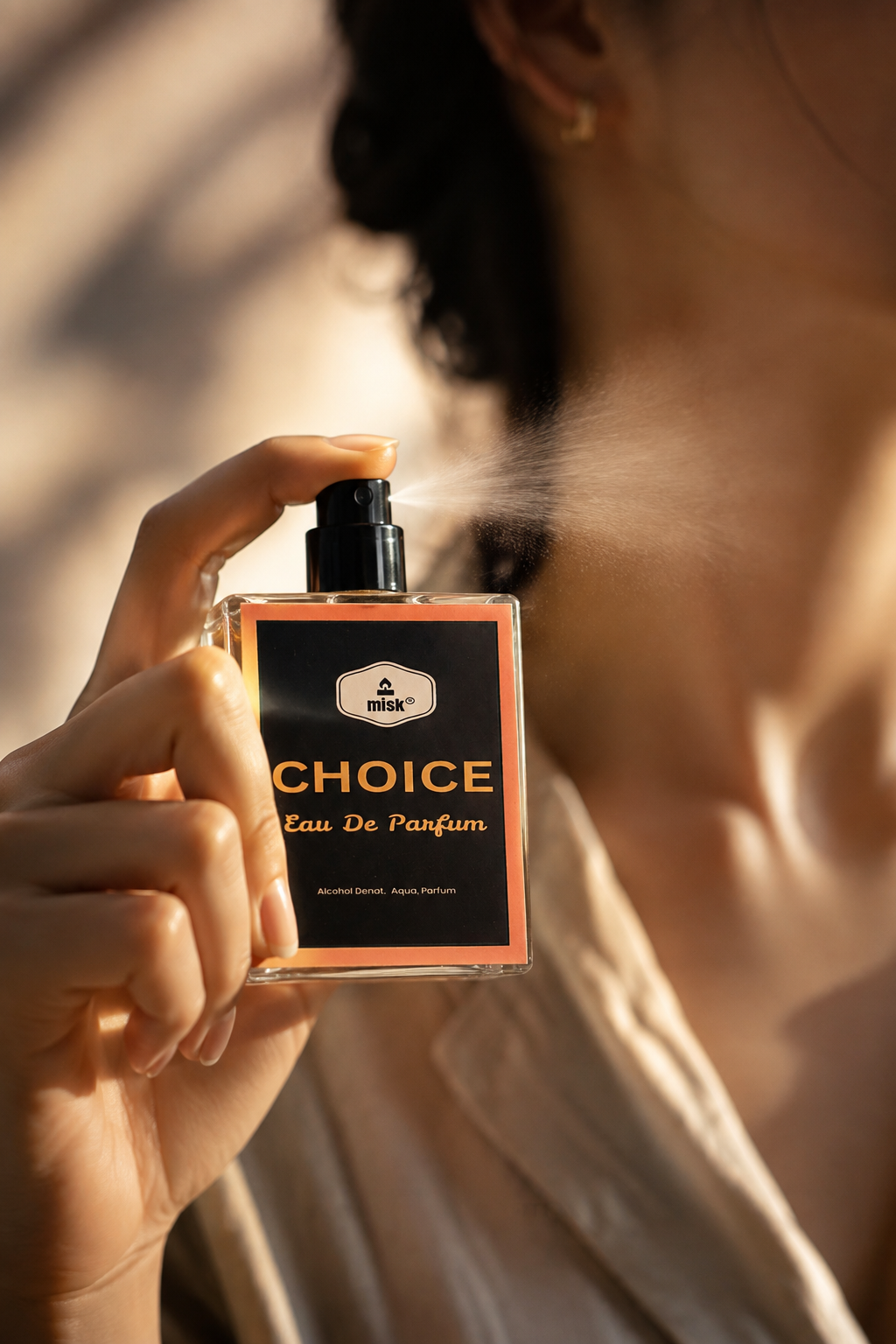 A person holding a bottle of CHOICE Eau De Parfum, spraying a fine mist. The person is wearing a light beige satin shirt, and the background is softly blurred with warm sunlight and shadows.