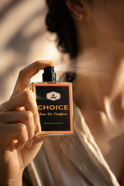 A person holding a bottle of CHOICE Eau De Parfum, spraying a fine mist. The person is wearing a light beige satin shirt, and the background is softly blurred with warm sunlight and shadows.