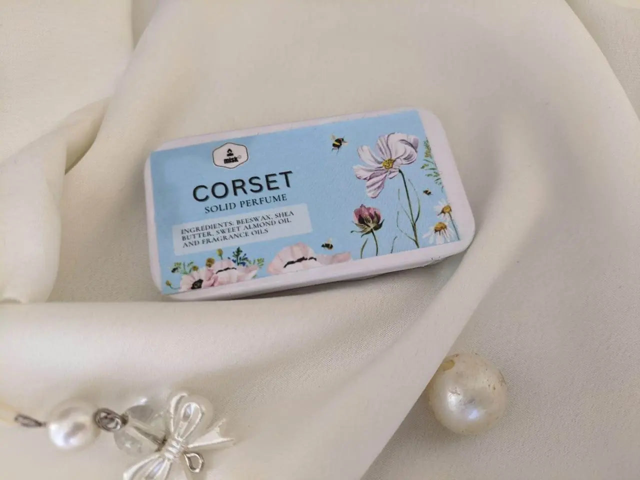 Solid perfume tin labeled 'Corset' on a white fabric background with decorative elements.