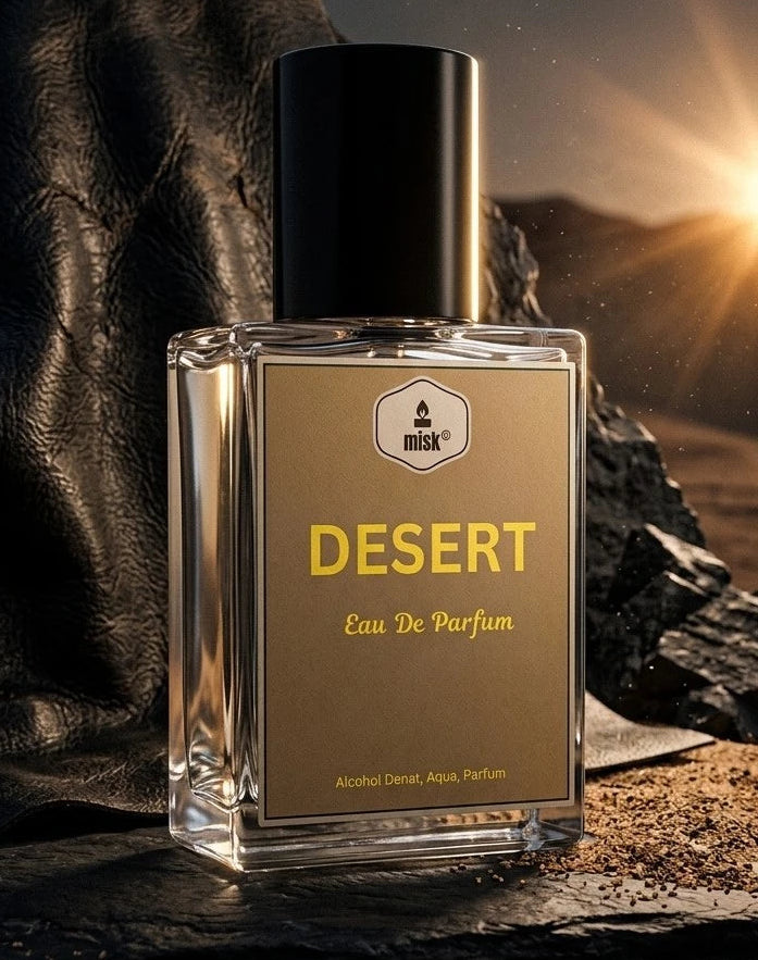 A rectangular glass perfume bottle labeled 'DESERT Eau De Parfum' with a black cap, set on dark rocks with a dramatic sunset glow in the background.