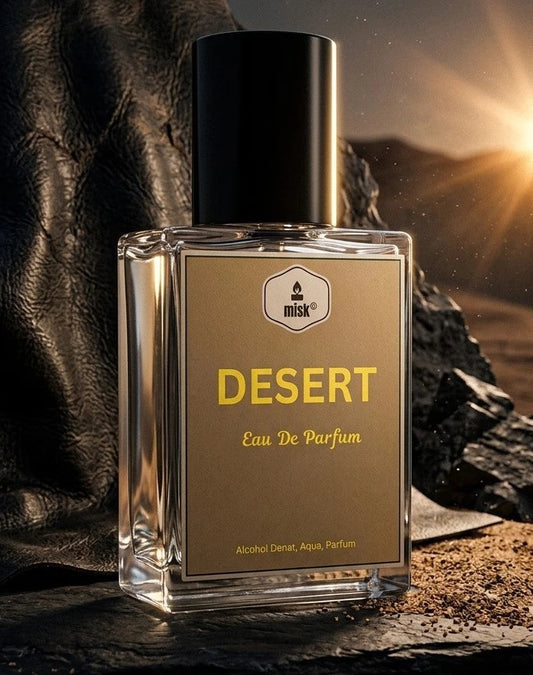 A rectangular glass perfume bottle labeled 'DESERT Eau De Parfum' with a black cap, set on dark rocks with a dramatic sunset glow in the background.