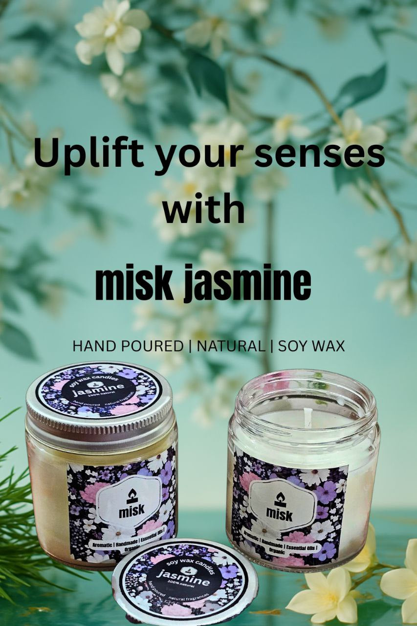 Two glass jars of Misk Jasmine soy wax candles with floral labels. One jar is closed with a lid showing "jasmine" text, and the other is open, revealing the white candle inside. The background features blurred white flowers and green foliage, with text overlay stating "Uplift your senses with misk jasmine" and "HAND POURED | NATURAL | SOY WAX".