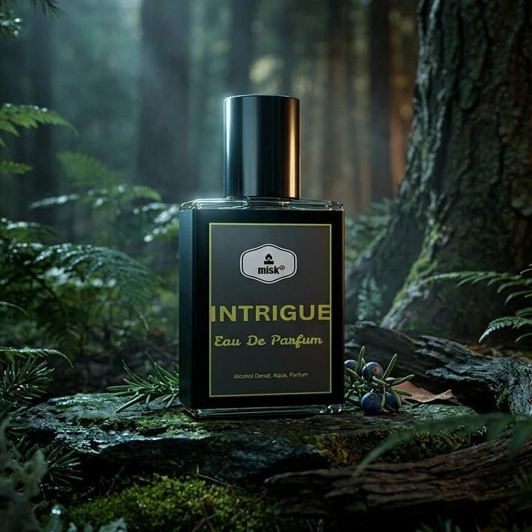 A rectangular perfume bottle labeled 'INTRIGUE Eau De Parfum' stands on a mossy log in a dark, misty forest with trees and ferns around it.