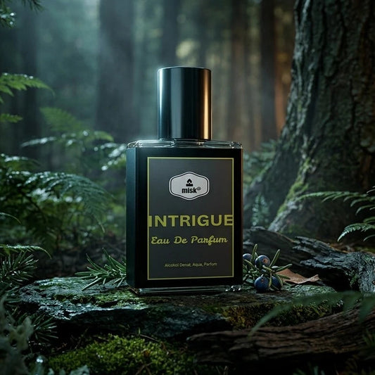 A rectangular perfume bottle labeled 'INTRIGUE Eau De Parfum' stands on a mossy log in a dark, misty forest with trees and ferns around it.
