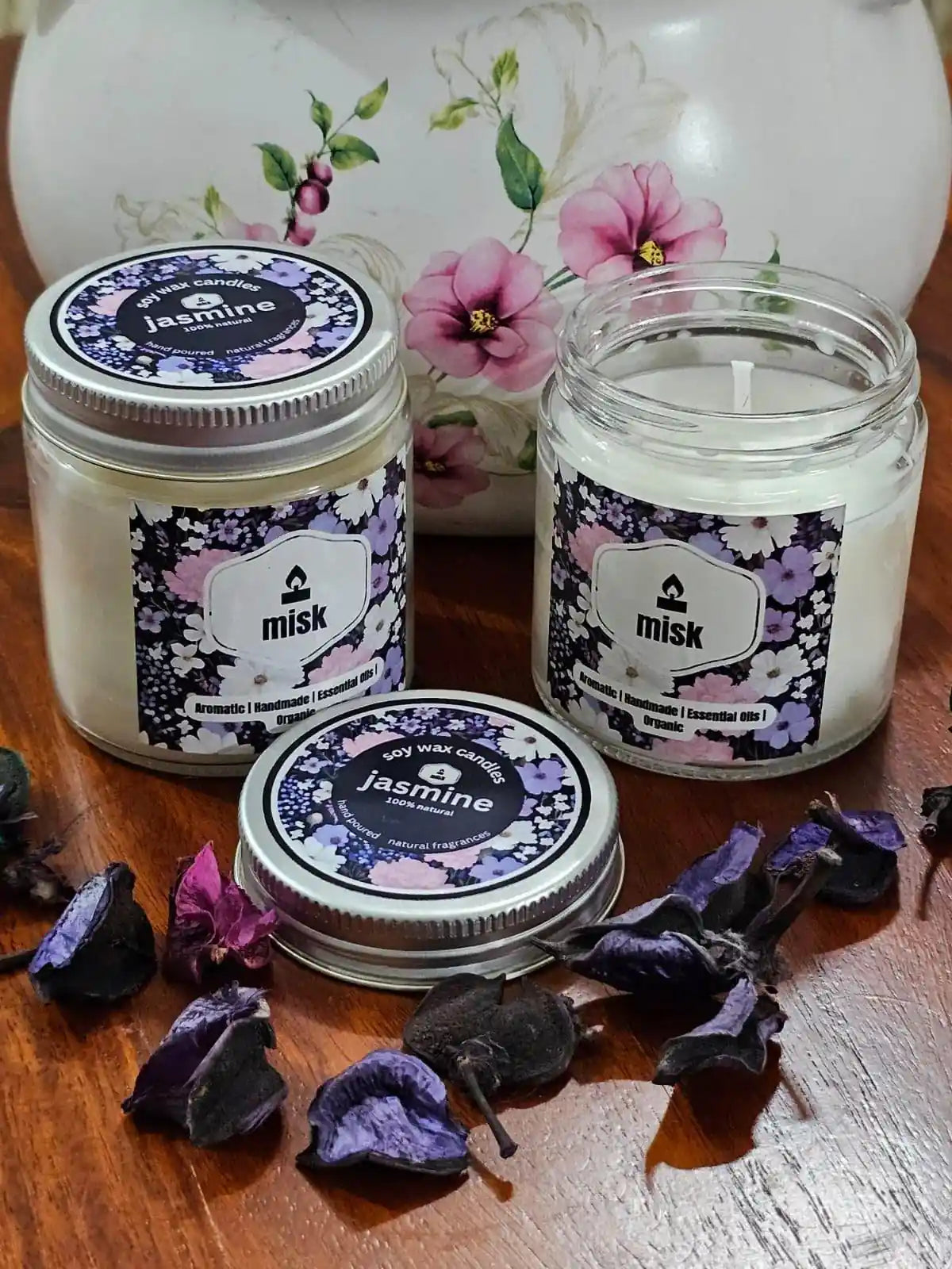Two jars of 'misk' candles with floral labels on a wooden surface with dried flowers.