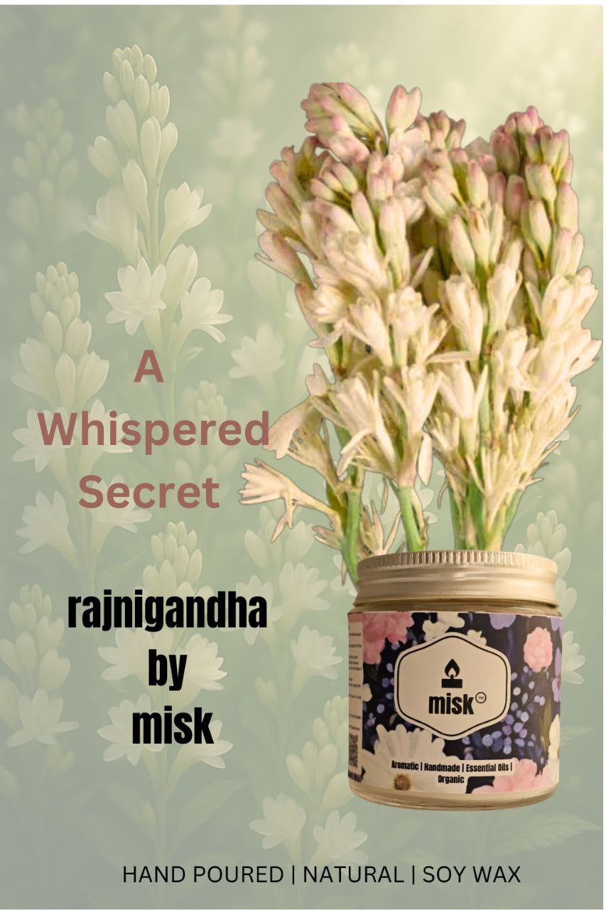 A close-up ad of a "misk" brand candle labeled "rajnigandha by misk" with text "A Whispered Secret," "HAND POURED | NATURAL | SOY WAX," and "Aromatic | Handmade | Essential Oils | Organic." The candle is placed next to a bouquet of white rajnigandha flowers, set against a blurred background of similar flowers.
