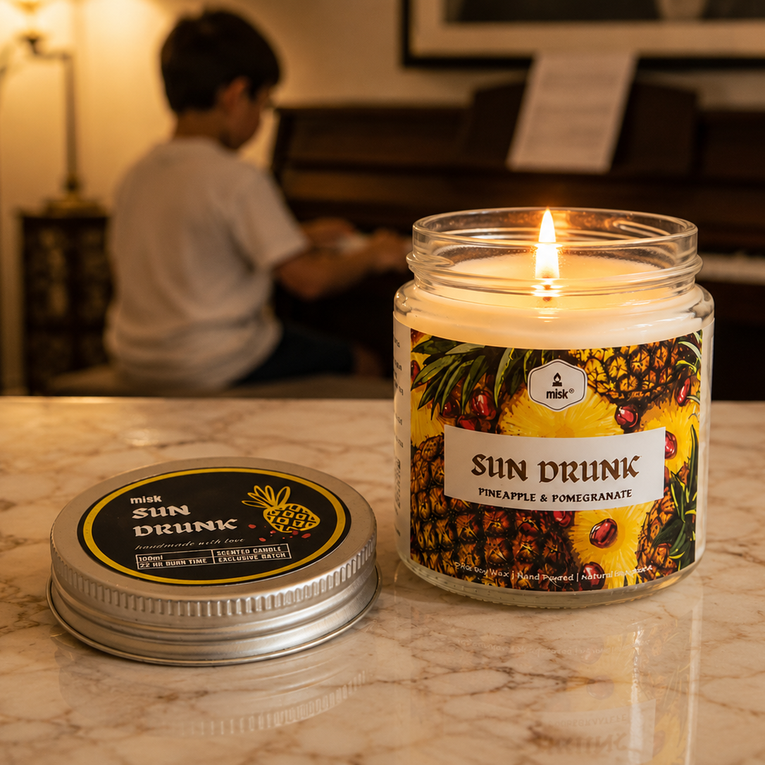 A burning Sun Drunk candle in the foreground on a marble top. A child playing a piano in the background