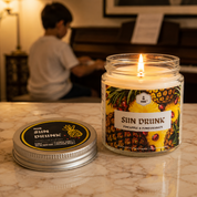 A burning Sun Drunk candle in the foreground on a marble top. A child playing a piano in the background